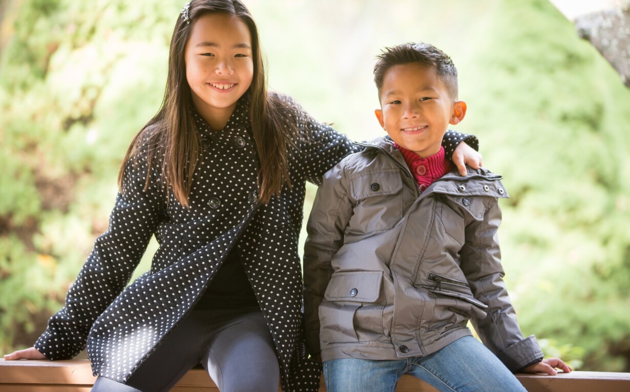 A young girl and boy sit on a wooden bench outdoors. The girl, in a black polka dot coat, has her arm around the boy in a gray jacket. They both smile at the camera amid lush greenery—a perfect picture of connection fostered by social emotional learning activities.