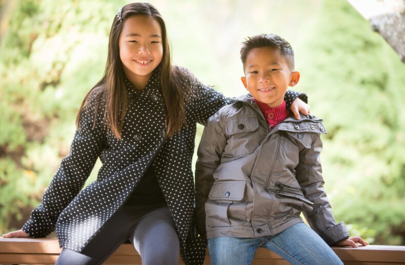 A young girl and boy sit on a wooden bench outdoors. The girl, in a black polka dot coat, has her arm around the boy in a gray jacket. They both smile at the camera amid lush greenery—a perfect picture of connection fostered by social emotional learning activities.