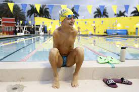 A smiling man wearing a yellow swim cap and goggles sits on the edge of an outdoor pool, embodying the essence of getting to know you activities. In swim trunks, with a water bottle, flip-flops, and a kickboard nearby, he's surrounded by palm trees and a bright yellow wall.