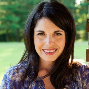 A woman with long dark hair smiles at the camera, standing outdoors in front of a blurred background with greenery. Wearing a patterned top, she appears in bright, natural light, embodying the positivity often celebrated in SEL activities that foster social connections.