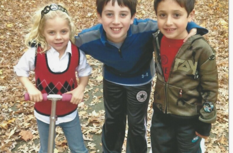 Three children are posing in a wooded area with autumn leaves on the ground, capturing a moment perfect for an SEL curriculum. The girl on the left holds a scooter, while the two boys stand next to her, smiling at the camera, embodying get-to-know-you activities for students in nature's classroom.