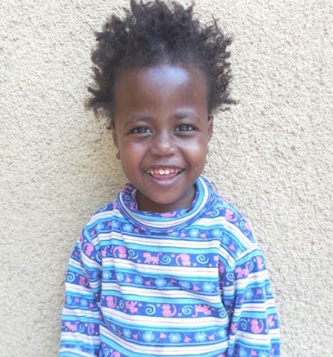 A smiling child with curly hair stands against a beige wall, wearing a colorful striped shirt, embodying the vibrant spirit of diversity programs.