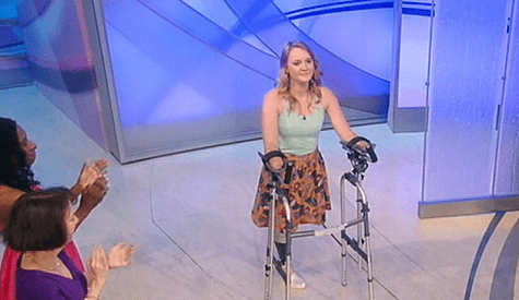A woman using a walker stands in the center of a room with two people clapping nearby, celebrating her spirit. She wears a light blue top and patterned skirt, embodying resilience. The setting, with modern abstract designs, feels like an extension of a social emotional learning curriculum in action.