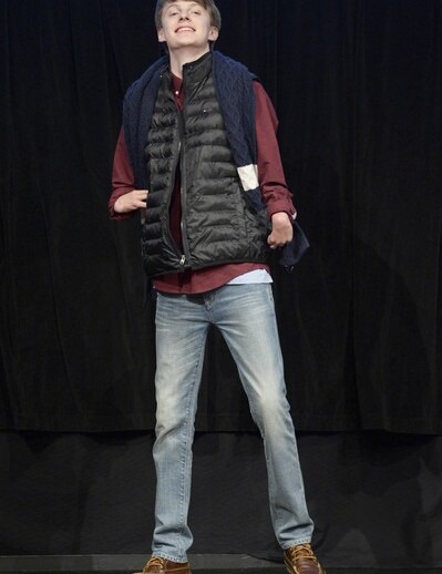 A person poses confidently on stage wearing a black puffy vest over a red shirt, with jeans and brown shoes, embodying the confidence-building focus of social emotional learning activities. A blue sweater is draped over their shoulders against a dark background.
