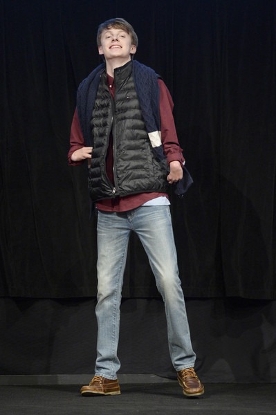 A person poses confidently on stage wearing a black puffy vest over a red shirt, with jeans and brown shoes, embodying the confidence-building focus of social emotional learning activities. A blue sweater is draped over their shoulders against a dark background.