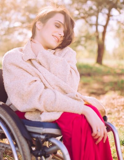 A woman sits in a wheelchair outdoors, wearing a beige sweater and red skirt. With a serene expression, she touches her neck amid the sunlit blur of trees and greenery—embracing the tranquility often celebrated in social emotional learning activities.