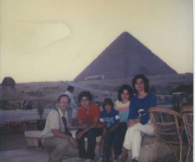 A group of five people poses together in front of a large pyramid at sunset, embodying the calm and relaxed atmosphere often found in social emotional learning activities. They sit and crouch on chairs and the ground, while a few tourists linger in the background.
