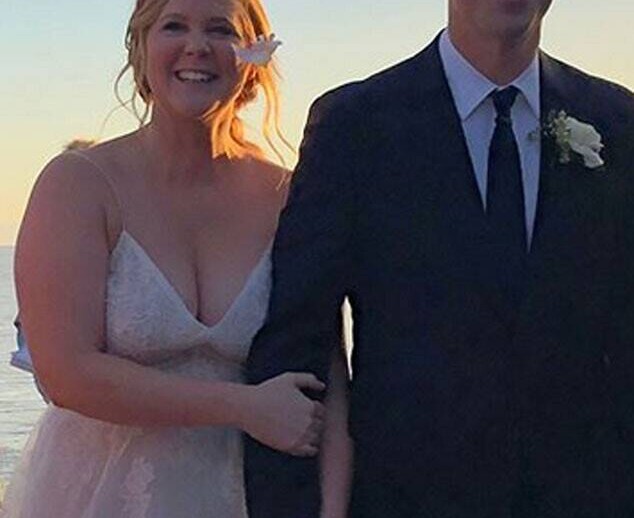 A bride and groom stand together at sunset, smiling warmly. The bride wears a white dress adorned with flowers in her hair, while the groom looks sharp in a black suit with a boutonnière. Amidst the partly cloudy sky's glow, their love blossoms like an SEL curriculum enriching young minds.