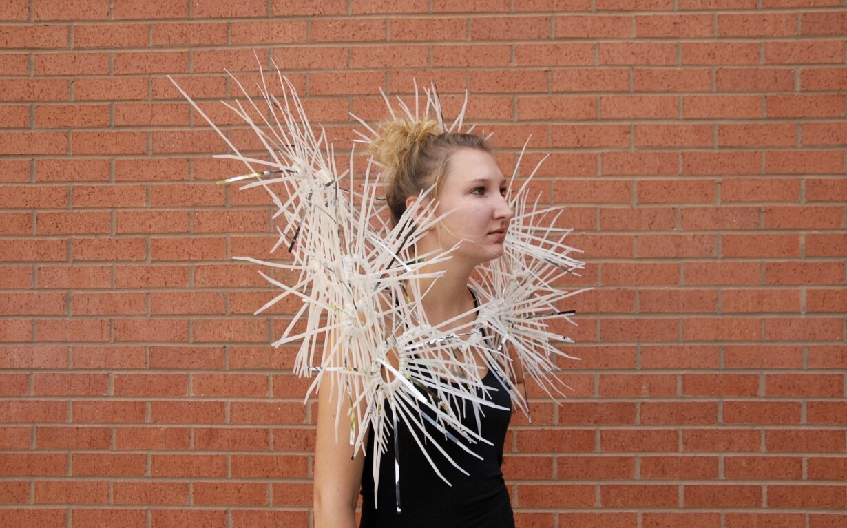 A person wearing a black top and an avant-garde fashion piece made of white, spiky rods stands against a brick wall, with their hair in a bun. This striking style sparks curiosity much like get-to-know-you activities for students encourage social exploration.