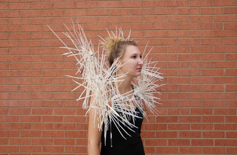 A person wearing a black top and an avant-garde fashion piece made of white, spiky rods stands against a brick wall, with their hair in a bun. This striking style sparks curiosity much like get-to-know-you activities for students encourage social exploration.