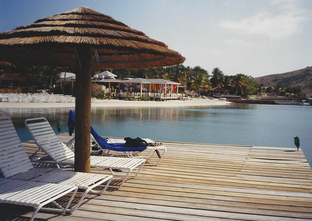 A wooden pier with several white lounge chairs under a thatched umbrella overlooks a calm bay, offering a serene setting perfect for SEL activities. Across the water, small buildings nestled among palm trees dot the sandy beach under a clear sky.