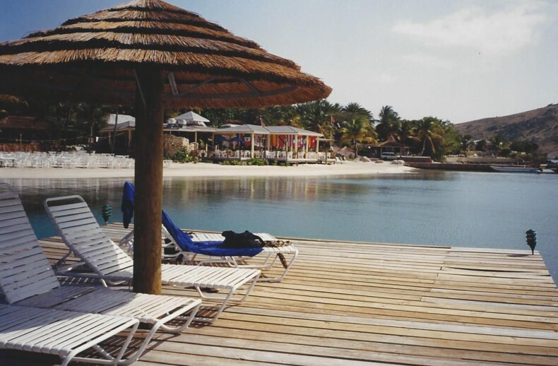 A wooden pier with several white lounge chairs under a thatched umbrella overlooks a calm bay, offering a serene setting perfect for SEL activities. Across the water, small buildings nestled among palm trees dot the sandy beach under a clear sky.