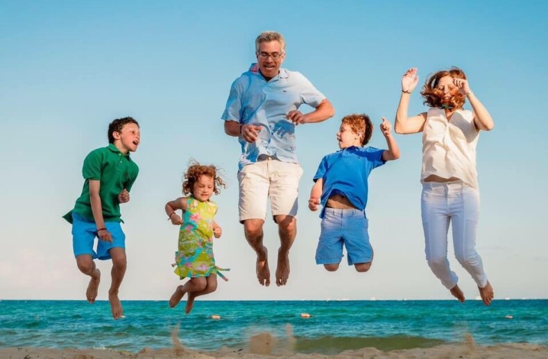 A family of five joyfully jumps in the air on a beach, much like kids during vibrant SEL activities. The sky is clear, and the sea is calm. Two children wear colorful summer clothes, while the adults are dressed casually. Everyone appears happy against the bright blue backdrop.