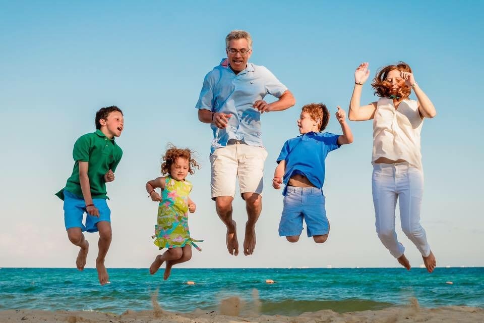 A family of five joyfully jumps in the air on a beach, much like kids during vibrant SEL activities. The sky is clear, and the sea is calm. Two children wear colorful summer clothes, while the adults are dressed casually. Everyone appears happy against the bright blue backdrop.