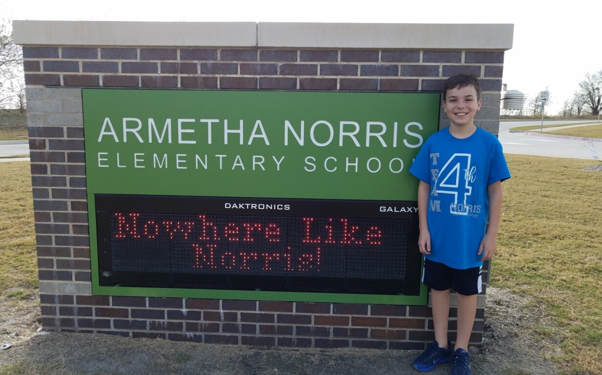A boy in a blue shirt stands smiling next to a brick sign reading "ARMETHA NORRIS ELEMENTARY SCHOOL" with a digital display that says "Nowhere Like Norris!" Perfect for enjoying SEL activities, the scene is outdoors on a sunny day.