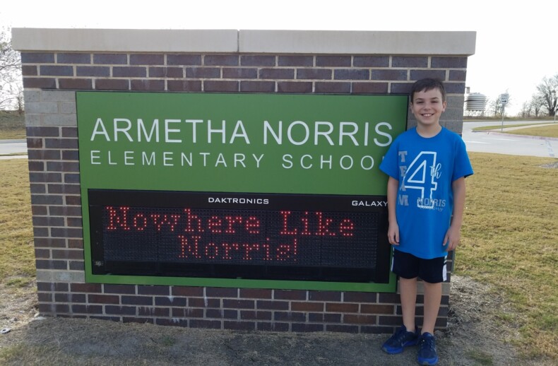 A boy in a blue shirt stands smiling next to a brick sign reading "ARMETHA NORRIS ELEMENTARY SCHOOL" with a digital display that says "Nowhere Like Norris!" Perfect for enjoying SEL activities, the scene is outdoors on a sunny day.