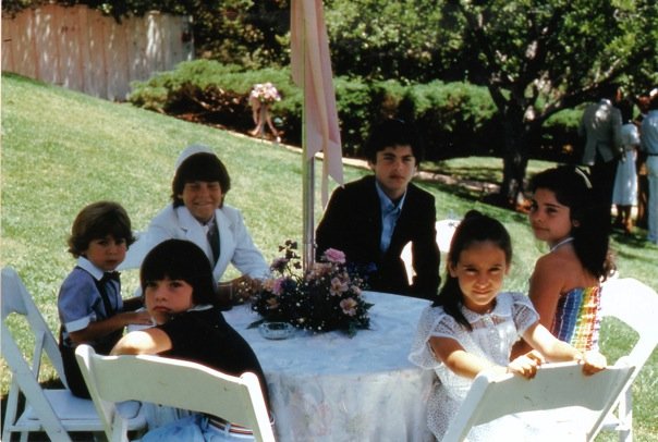 A group of young people sit around a table outdoors in a garden setting, engaging in SEL activities. The table is covered with a white cloth and a floral centerpiece. Trees and bushes can be seen in the background on this sunny day, adding to the serene atmosphere.