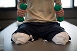 A person, possibly engaged in social emotional learning activities, is sitting on a padded surface, holding two green dumbbells. They appear to be wearing a beige shirt and dark shorts over white padded areas on their knees. The background is softly lit by window light.
