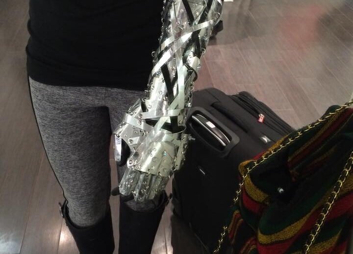 A person is standing indoors wearing a stylish outfit with gray leggings and black boots. Their forearm is wrapped in a creation made of clear plastic and metallic strips, an emblem of their focus on SEL curriculum. They're surrounded by a suitcase and a multicolored bag.