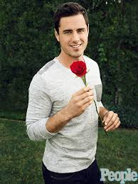 A man in a gray sweater holds a red rose while standing on a lawn, embodying themes of diversity from social emotional learning curriculum. Bushes form the backdrop, and a magazine logo is visible in the bottom right corner.