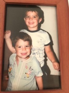 Two young children smiling, with the older one standing playfully behind the younger, embodying the joy of social-emotional learning activities. Both are in casual clothes, captured beautifully in a wooden frame that celebrates their diverse friendship.