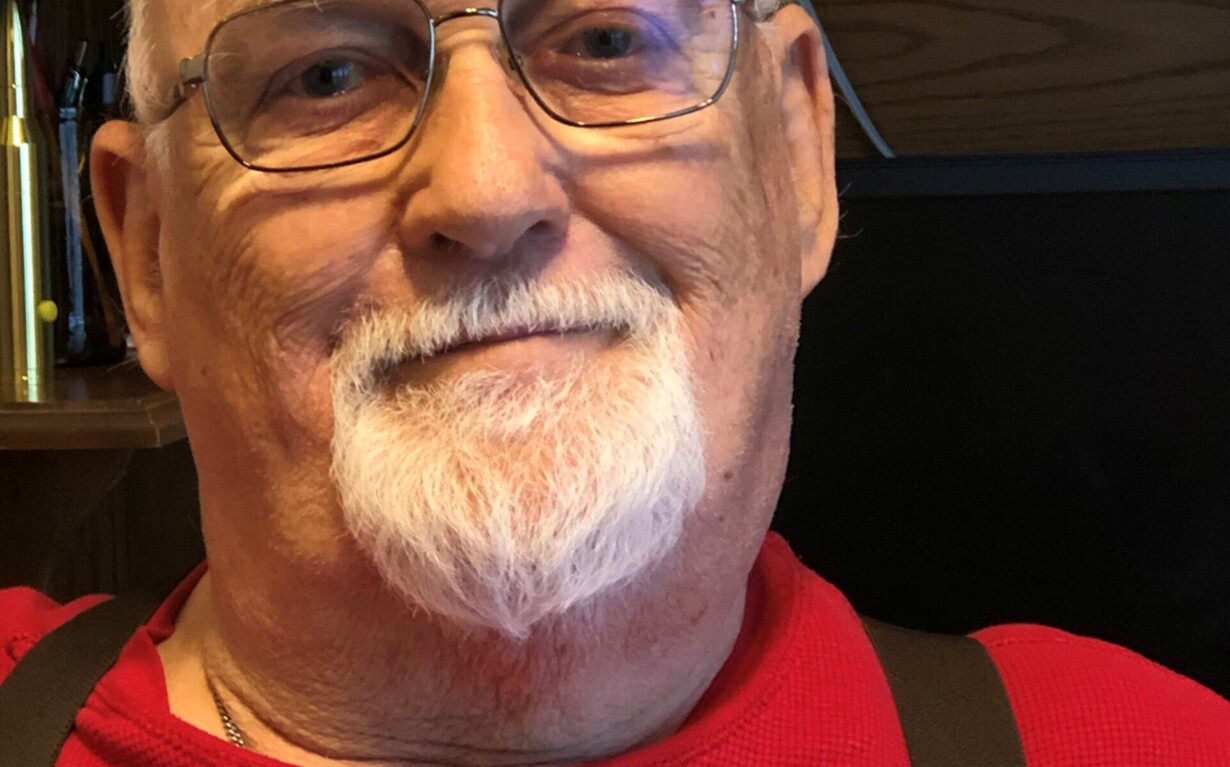 An elderly man with a white beard and glasses smiles slightly while wearing a red shirt and suspenders. Seated indoors with wooden furniture in the background, he exudes warmth—a perfect presence for engaging in social emotional learning activities that bridge generations.