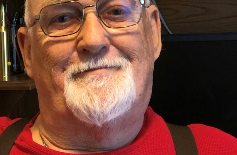 An elderly man with a white beard and glasses smiles slightly while wearing a red shirt and suspenders. Seated indoors with wooden furniture in the background, he exudes warmth—a perfect presence for engaging in social emotional learning activities that bridge generations.