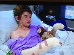 A young person with short brown hair lies in a hospital bed, wearing a purple gown. Bandages cover their arms as they clutch a small stuffed lion. Medical equipment surrounds them, while thoughts of SEL activities bring comfort during this challenging time.
