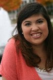 A woman with long brown hair smiles at the camera, wearing a peach-colored top and pearl necklace. The background features a blurry outdoor setting with autumn-colored trees and a fountain, hinting at serene moments often found in social emotional learning activities.