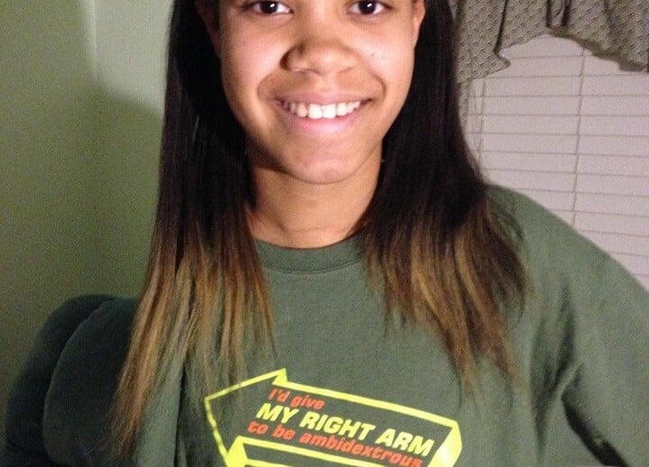 Smiling person with long hair wearing a green shirt that humorously promotes social emotional learning. The background showcases a room with a window and curtains, reflecting an atmosphere enriched by diversity programs.