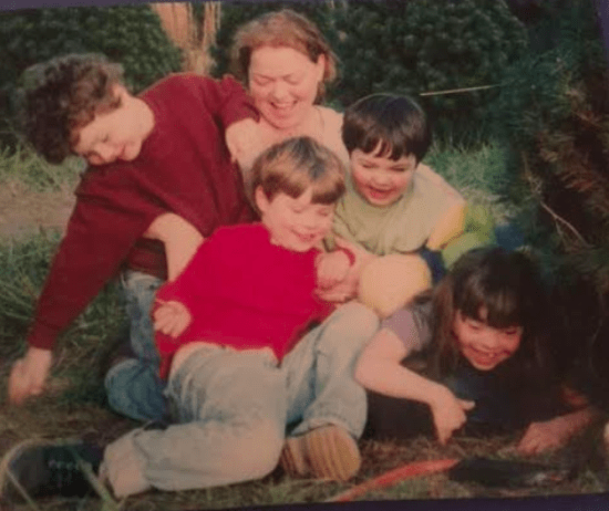 A group of children and an adult laugh and play on the grass among trees, engaging in fun SEL activities. The children are wearing colorful sweaters, and everyone appears joyful and relaxed, enjoying a playful moment outdoors together.