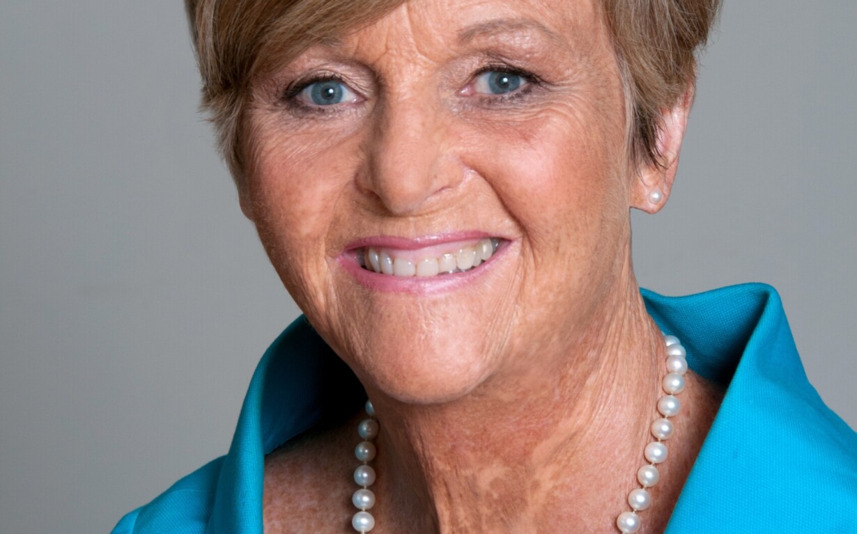 A smiling elderly woman with short hair, wearing a bright blue jacket and a pearl necklace, embodies the warmth and inclusivity championed by diversity programs as she poses against a plain background.