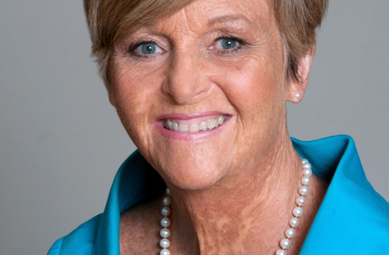 A smiling elderly woman with short hair, wearing a bright blue jacket and a pearl necklace, embodies the warmth and inclusivity championed by diversity programs as she poses against a plain background.