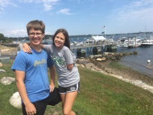 Two people are smiling, standing on a grassy area near a rocky shoreline. Behind them is a dock with several boats anchored on a sunny day. By embracing social emotional learning curriculum, they've found joy and connection under the partly cloudy sky.