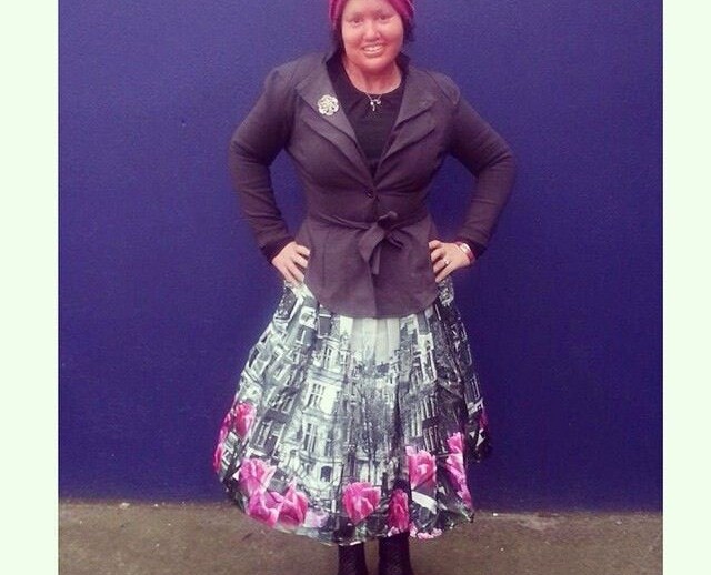 A person stands against a blue wall wearing a black jacket, a skirt with building and floral patterns, black boots, and a red headdress. Embracing diversity programs, they smile with hands on hips, radiating confidence and celebrating individuality in style.