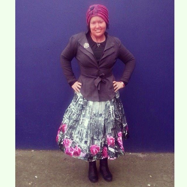 A person stands against a blue wall wearing a black jacket, a skirt with building and floral patterns, black boots, and a red headdress. Embracing diversity programs, they smile with hands on hips, radiating confidence and celebrating individuality in style.