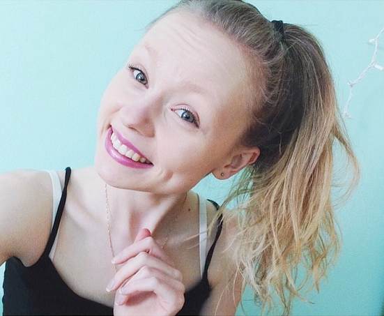 A smiling person with their hair tied back in a ponytail poses in front of a light blue background, embodying the spirit of diversity programs. They are wearing a black top and a necklace.