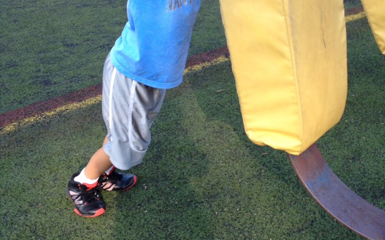 A young boy in a blue shirt and gray shorts playfully interacts with a large yellow football tackling dummy on a grassy field. As part of SEL activities, other people and sports equipment create an engaging backdrop, fostering teamwork and emotional growth.