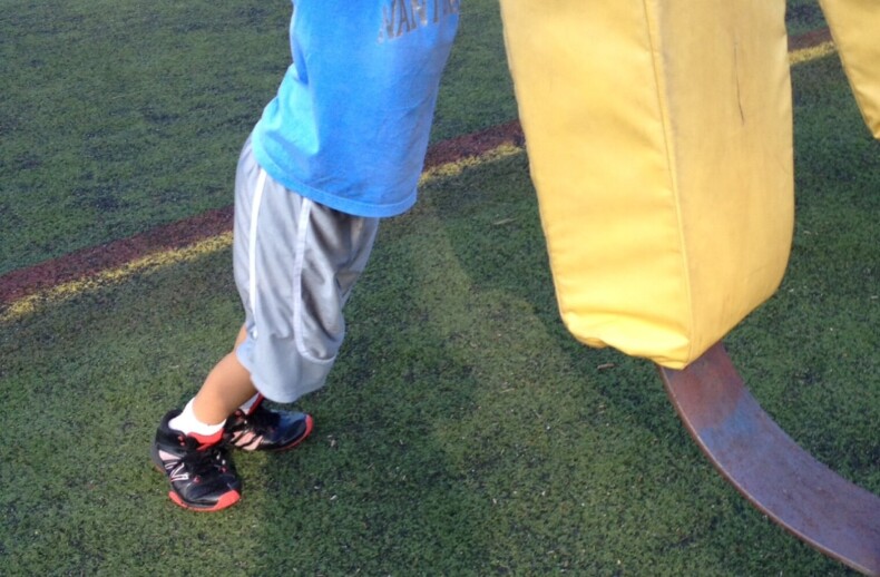 A young boy in a blue shirt and gray shorts playfully interacts with a large yellow football tackling dummy on a grassy field. As part of SEL activities, other people and sports equipment create an engaging backdrop, fostering teamwork and emotional growth.
