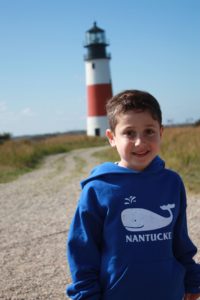A boy in a blue hoodie featuring a whale and the word "Nantucket" stands smiling on a gravel path. Behind him, a red and white striped lighthouse is visible under a clear blue sky—the perfect setting for engaging SEL activities that build empathy and connection.
