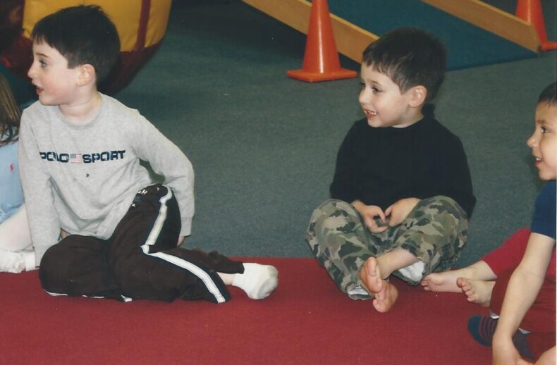 Three young boys sit on a red carpet in a play area, immersed in SEL activities. One wears a gray shirt, another sports a black top with camouflage pants, and the third dons a red shirt. Barefoot and engaged, they explore colorful equipment that promotes growth and understanding.