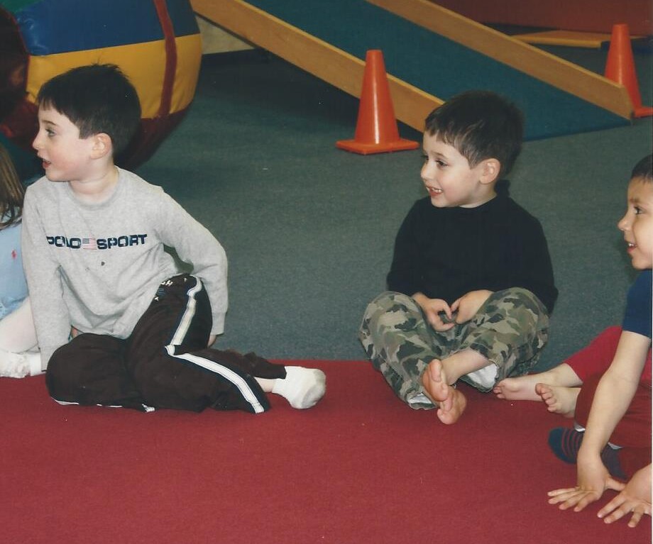 Three young boys sit on a red carpet in a play area, immersed in SEL activities. One wears a gray shirt, another sports a black top with camouflage pants, and the third dons a red shirt. Barefoot and engaged, they explore colorful equipment that promotes growth and understanding.