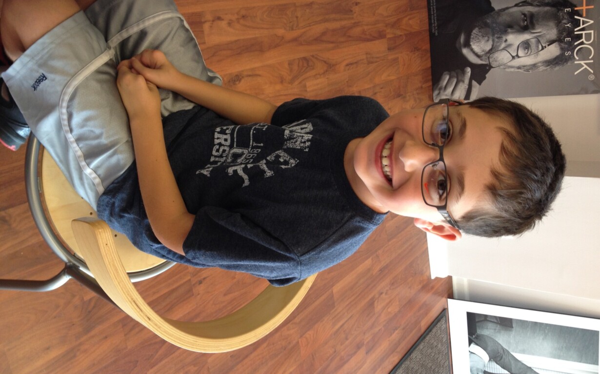 A young boy wearing glasses sits on a wooden chair, smiling at the camera after a fun social emotional learning activity. He wears a dark t-shirt and light shorts, with a poster of an older person with glasses behind him against the wooden floor backdrop.