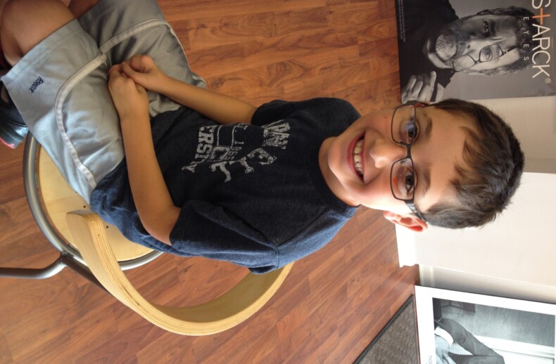 A young boy wearing glasses sits on a wooden chair, smiling at the camera after a fun social emotional learning activity. He wears a dark t-shirt and light shorts, with a poster of an older person with glasses behind him against the wooden floor backdrop.