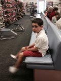 A young boy in a white shirt and red shorts sits on a blue bench in a shoe store, exploring get-to-know-you activities for students on his tablet. He seems to be waiting, surrounded by shelves of shoes and other customers bustling about.