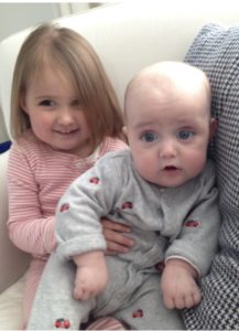 A young child with shoulder-length hair, wearing a pink-striped outfit, is sitting on a couch holding a baby dressed in light grey pajamas. The baby has big eyes and a curious expression, embodying the curiosity central to SEL activities. Both are seated on a patterned pillow.