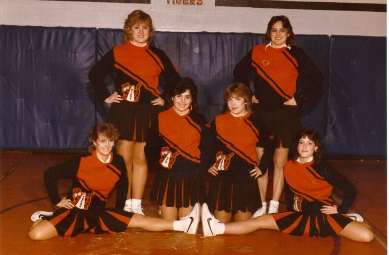 Six cheerleaders in black and orange uniforms pose confidently in a gymnasium, reflecting the energy of their social emotional learning curriculum. Four stand with hands on hips, while two sit cross-legged in front. The word "TIGERS" proudly adorns the wall behind them.