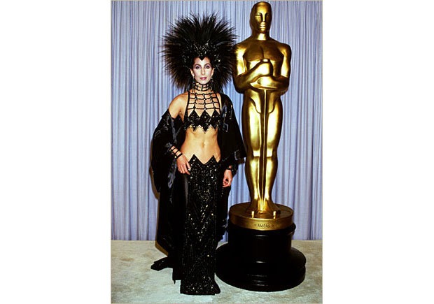 A person wearing an elaborate black outfit with a headpiece stands beside a large golden statue in front of a curtain backdrop, embodying the essence of diversity programs.