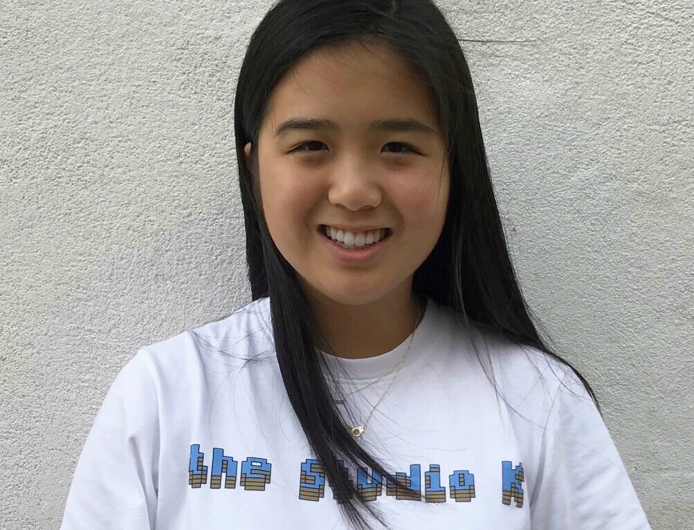 A person with long dark hair is smiling while standing against a light-colored wall, embodying the warmth often found in social emotional learning curriculum. They are wearing a white T-shirt with blue text on it.
