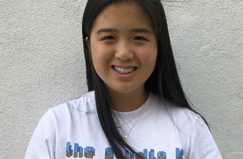 A person with long dark hair is smiling while standing against a light-colored wall, embodying the warmth often found in social emotional learning curriculum. They are wearing a white T-shirt with blue text on it.